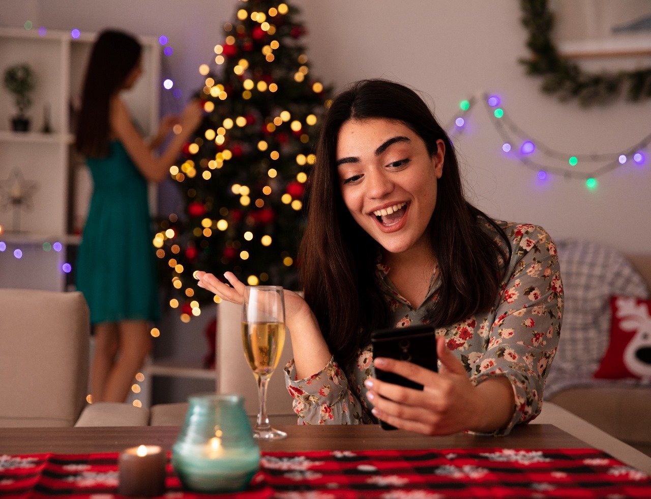 pleased-pretty-young-girl-decorates-christmas-tree-her-surprised-friend-looks-phone-sitting-table-enjoying-christmas-time-home